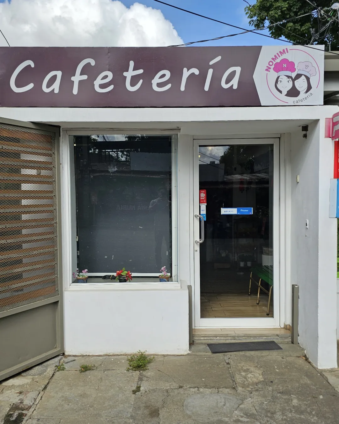 Fachada de Nomimi: letrero color vino que dice Cafetería, con el logo de las hermanas, y la puerta de vidrio